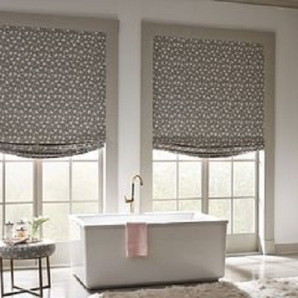 Roman shades on windows behind a white bathtub.